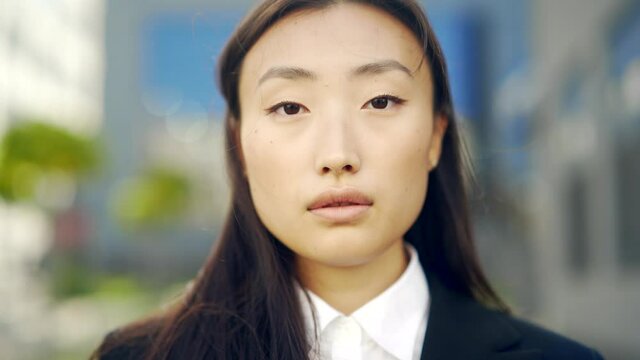 close-up portrait of young attractive asian business woman looking at camera on urban city street background. face of independent confident businesswoman or student. outside. outdoors. Closeup