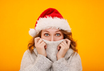 Happy young woman in white winter sweater wearing santa claus hat and hiding her face isolated on yellow orange background.