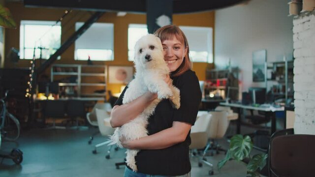 Young Beautiful Caucasian Woman Holding an Adorable Dog and Giving a Gentle Kiss in an Industrial and Modern Office Space. Colorful Background Filled With Mordern Interior Pieces.