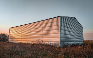 Fototapeta na wymiar Warehouse constructed of corrugated galvanized iron. Warehouse in autumn during sunset in a field with dead vegetation.