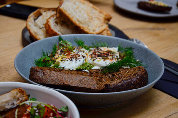 On the table in a plate is a slice of rye bread, poached eggs sprinkled with cereal seeds and lined with dill sprigs. Behind her is a plate of toasted white bread.