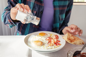 Sprinkles eggs and bacon with salt from a shaker. An excess of sodium chloride mineral can lead to...