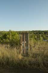 A strange shabby wooden door in the middle of the road against the background of bushes, green deciduous forest and blue clear cloudless sky in summer