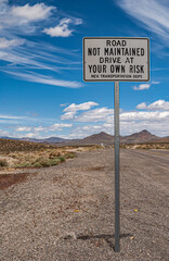 Tonopah, Nevada, US - May 16, 2011: Closeup of road sign warning road is not maintained, drive at your own risk against blue cloudscape. Gray mountains in back.