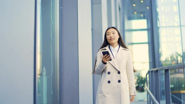 Business woman or university college student in a suit walks down a city street, talks and uses a mobile phone, smartphone. Asian young female lady office worker employee In a trench coat