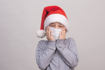 Fototapeta na wymiar A boy in a Santa helper hat and a medical protective mask covers his face with his hands. The child is offended and upset in stress, wants to cry. on a white background.