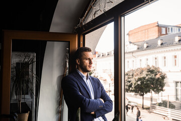 Handsome male model is posing in cafe. Bearded man weared suit stand and smile near window.