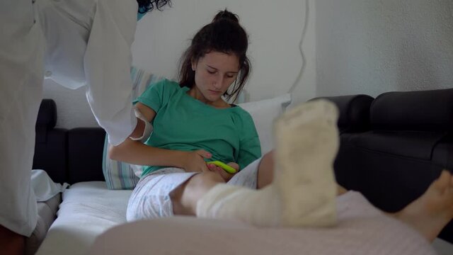 Young woman with broken leg is laying on couch sofa, resting and using her smartphone. Doctor helping her with a pillow