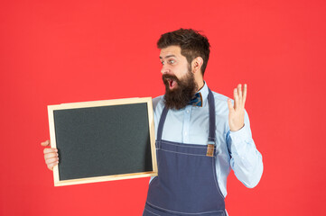 Surprised bearded man in barber apron hold school blackboard red background copy space, enrollment