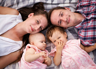 Fototapeta premium Happy family lies on a picnic blanket together - Young parents with a toddler and a baby daughter having fun - Bonding, love and care lifestyle concept from top view