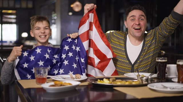 Excited Caucasian man and teenage boy rejoicing shaking American flag dining in restaurant watching football match. Portrait of happy smiling sports fans gesturing victory in slow motion. Father son