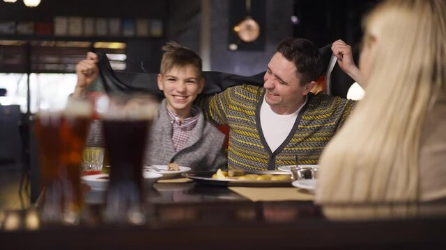 Portrait of smiling excited happy father and son hugging shaking German flag watching sport match in restaurant with wife mother. Thrilled Caucasian man and teenage boy rejoicing winning