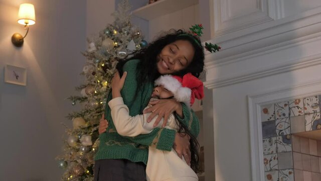 African american mom in green sweater and headband with christmas trees hugs her little daughter in a christmas hat against the background of a christmas tree.