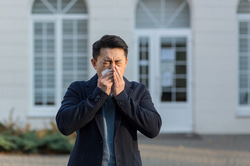 An Asian sick man sneezes near the office on the street, has allergies and illness