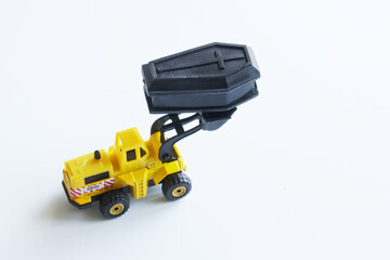Yellow toy forklift is holding a black toy coffin in a bucket. Concept of the exhumation coffin and...