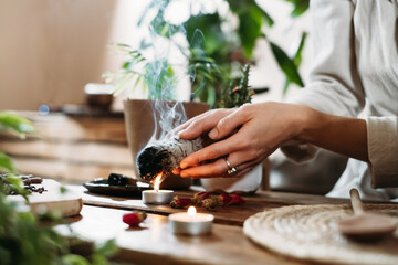 Woman hands burning white sage, palo santo before ritual on the table with candles and green...