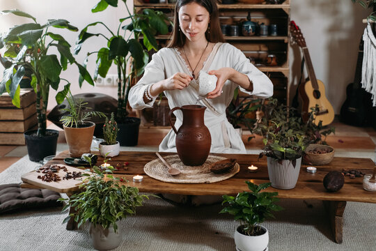 Pouring water into jug with cacao. Preparing ceremonial cocoa in atmospheric boho style cafe full of plants. Woman cooking healthy drink from organic cacao beans for ritual 