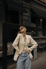 Young beautiful blonde posing effectively with her head thrown back, holding her hands in jeans pockets. Girl in black glasses and beige jacket stands against backdrop of dark building of street.