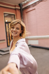Close-up of happy caucasian young blonde woman laughs and stretches her hand to camera while on street. Short-haired lady with tousled hair has fun smiling broadly in photo,