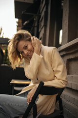 Cheerful caucasian young beauty with short hair straightens her hair behind ear in open space. Girl sits on chair, smiling broadly with her teeth at camera, wearing casual autumn clothes.