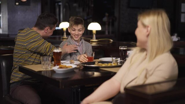 Rack focus joyful father and teenage son having fun talking smiling sitting in restaurant to embarrassed woman at front looking at camera with dissatisfied facial expression. Individuality concept