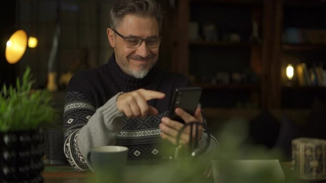 Older man sitting at desk in dark cosy room working with phone in home office. Mature age, middle age, mid adult casual man in 50s, confident happy smiling.
