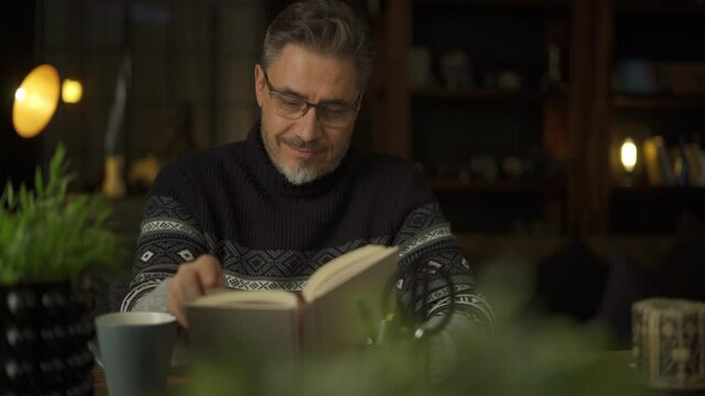 Older man sitting at desk in dark room reading book. Mature age, middle age, mid adult casual man in 50s, confident happy smiling. Cosy winter evening.