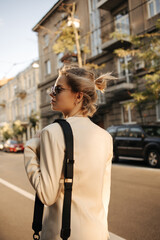 Fototapeta premium Rear view of stylish fair-haired girl in black glasses walking along city street looking to side. Her short hair is pulled back in bun, wearing white cashmere jacket and shoulder bag.
