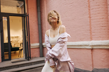 Caucasian young woman stands smiling broadly against background of pink house. Her short blonde hair blows in wind, covering her face. Dressed in white top, shirt and pants.