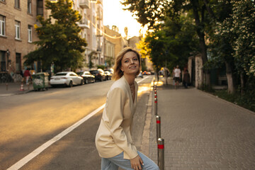 Fototapeta premium Not full-length caucasian young woman cute smiling on street. Girl with short hair crosses road, looking around in beige jacket and light-colored jeans. Warm day for walk in fresh air.