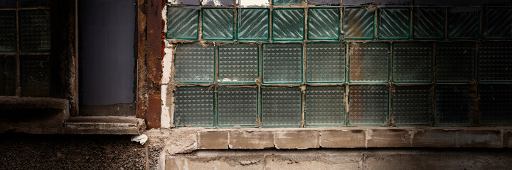 Old wall with a window made of square gray and glass tiles. Rough ribbed surface of dirty green glass. Vintage texture for background and design.