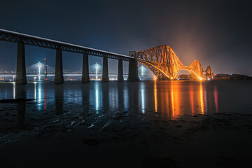 View of Forth Rail Bridge at night and and the glow trail of a moving train over the sea. View of...