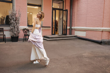 Amazing slim caucasian young blonde posing sideways in full growth against backdrop of street cafe. Girl with bob haircut turns around looking at camera, dressed in casual white clothes.