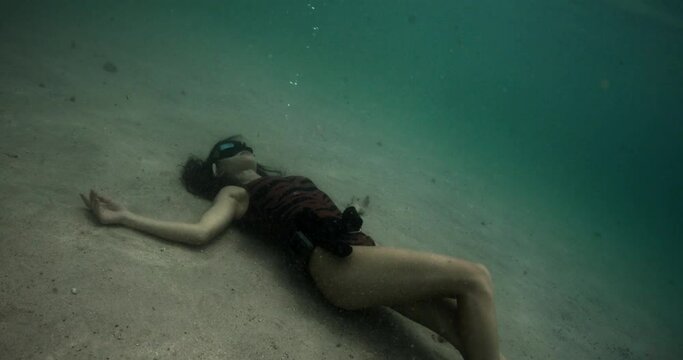 Freediver woman lying underwater bottom, filmed on cinema camera. Brutal girl in black suits against background of mountains and ocean. Beautiful brunette touching her shoulder. Concept of arthouse