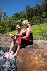 smiling couple sitting on the rock, wetting feet in river water in sunny day.