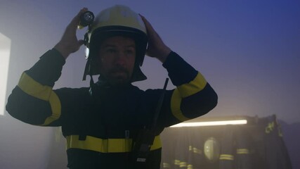 Mature caucasian firefighter putting on helmet indoors in fire station, getting ready for action at night