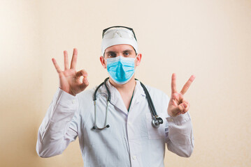 Present doctor in white suit and protective breathing mask glasses and stethoscope showing peace signs V and OK by his hands