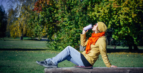 Naklejka premium Pretty european woman in yellow jacket and red scarf sitting on wooden bench in a deserted park and drinking coffee. Enjoying solitude and sunny autumn weather. Blurred plant background, copy space.