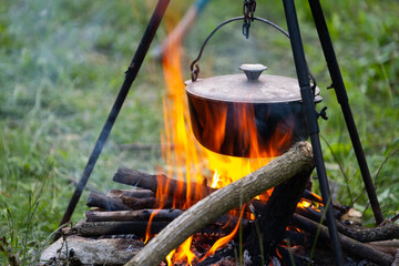 Black large tourist pot hangs over burning flame of campfire in the forest. Cooking dinner on wild...