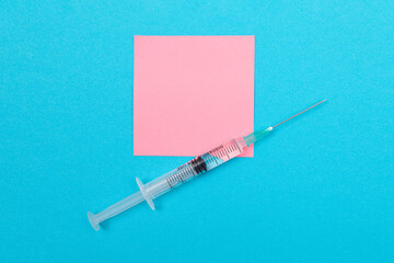 Vaccination, Immunology or Revaccination Concept - A Medical Disposable Syringe Lying on Blue Table in Doctor's Office in a Hospital or Clinic. Blank Pink Sticky Note - Mock Up with Copy Space
