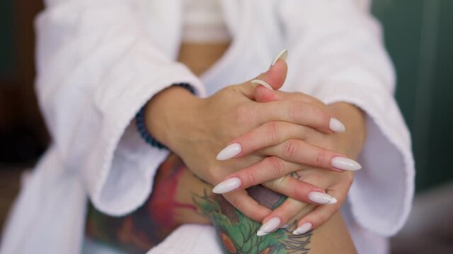 Woman with manicure and tattoos on leg in gown sits in room