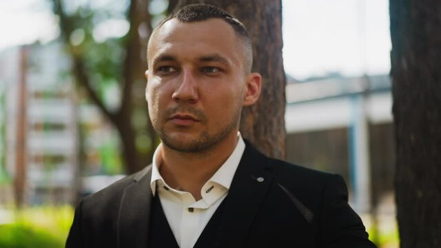 Man with short haircut and bald patches wearing classic black suit and white shirt with unbuttoned collar looks aside standing in city park closeup slow motion