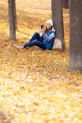 Video call outdoor. Woman in blue warm wear sits under a tree and greeting someone using mobile in the park in golden autumn, vertical photo, selected focus.