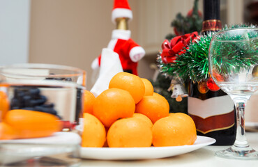 On the festive table there is a plate with tangerines, a bottle of champagne, New Year and Christmas decor