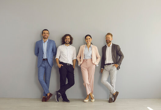 Group Portrait Of Four Successful Young Business People In Formal And Smart Casual Wear. Team Of 4 Happy Colleagues Or Professional Executive Managers Standing Together Near Grey Studio Or Office Wall