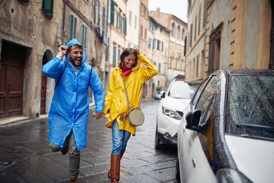 A young cheerful couple in raincoats is running from the rain towards the car while took a walk the city. Walk, rain, city, relationship
