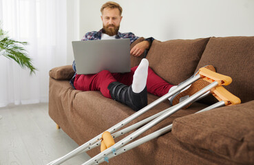 Serious man with broken injured leg sitting on sofa with crutches at home, using laptop computer...