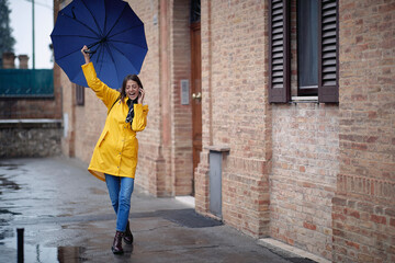A young handsome woman in a yellow raincoat and with umbrella is having a phone call while walking the city on a rainy day. Walk, rain, city