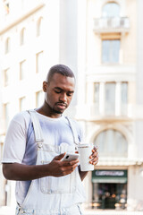 afro man walking the streets and using his smart phone. He is dressing casual summer clothes and holds an reusable cup of coffee