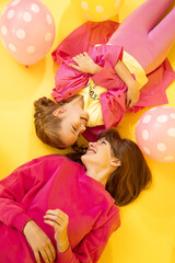 2 girls sisters are lying on yellow background in studio looking at each other and smiling, balloons, happy concept of childhood, relationship between children in family. Holiday, child psychology